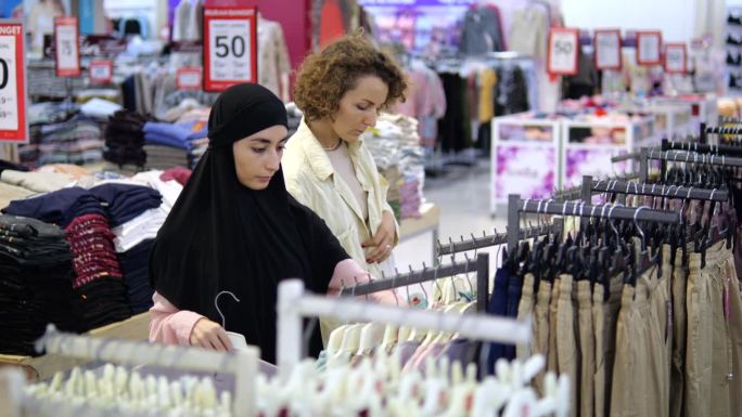 两位顾客在女装店里挑选东西、购物。戴着头巾的穆斯林妇女在精品店看衬衫、t恤和衬衫。不同国籍的顾客在店