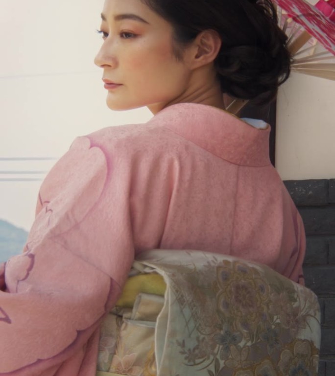 垂直屏幕:美丽的日本模特在一个广告活动中穿着粉红色和服摆姿势。年轻迷人的女性站在房子外面，拿着一把传