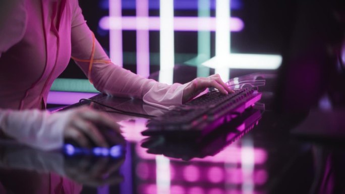 一个女玩家女孩在电脑上玩游戏的特写。专注于使用键盘和鼠标的手。坐在彩色霓虹灯工作室的未来互动桌子后面