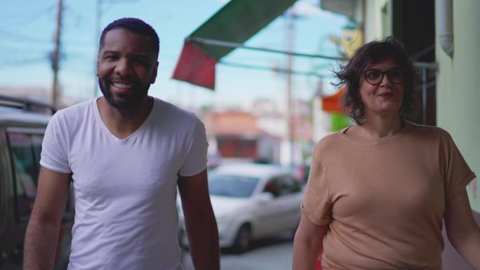 两个不同的巴西人慢镜头走在城市街道的人行道上。一个快乐的黑人男子向前走，旁边是一个成熟的中年妇女