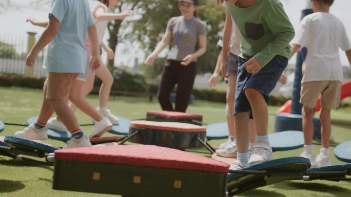 在温暖的阳光下，欢快的老师和来自不同国家的幼稚园小朋友们在小学的绿色公园里练习平衡。