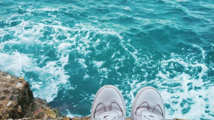 照片中是穿着白色潮鞋的徒步旅行者的双脚。蔚蓝的海浪或海洋的壮丽景色，度假游客的脚位于岩石上。