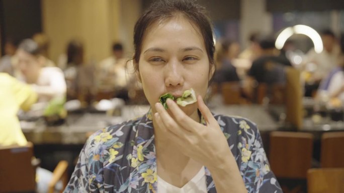 一位快乐的亚洲妇女在一家拥挤的日本烧烤店和朋友一起吃午餐时，用筷子和手咬了一大口生菜和烤猪肉，并竖起