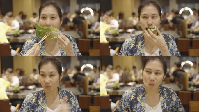 一位快乐的亚洲妇女在一家拥挤的日本烧烤店和朋友一起吃午餐时，用筷子和手咬了一大口生菜和烤猪肉，并竖起