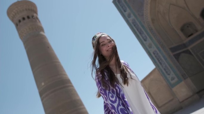 一位身穿白色连衣裙和东方长袍的年轻女子在阳光下跳舞，旁边是一座古老的尖塔