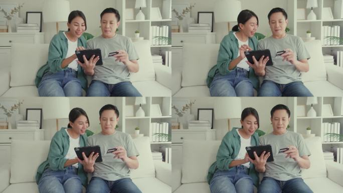 幸福的亚洲夫妇一起坐在家里客厅的沙发上玩电子平板设备购物，使用信用卡在线支付。生活方式花时间在家里。