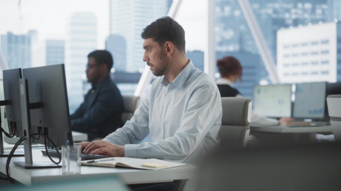 快乐的男性客户经理在不同公司办公室的台式电脑上打字，拥有大都市的窗口视图。专业的白种人为咨询公司做笔