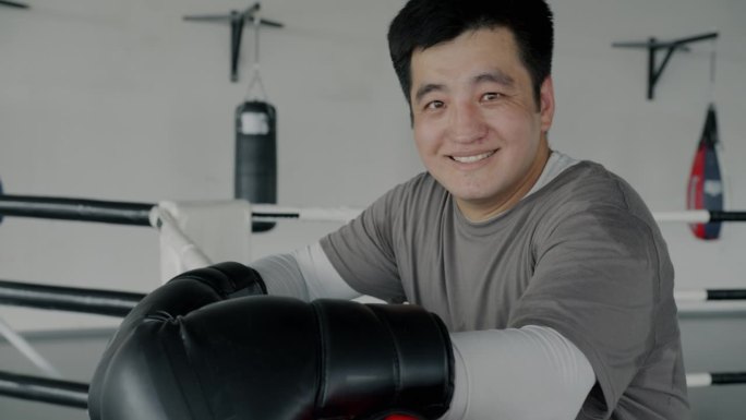 快乐的亚洲男子拳击手站在拳击场上，微笑着看着镜头，享受在健身房的训练