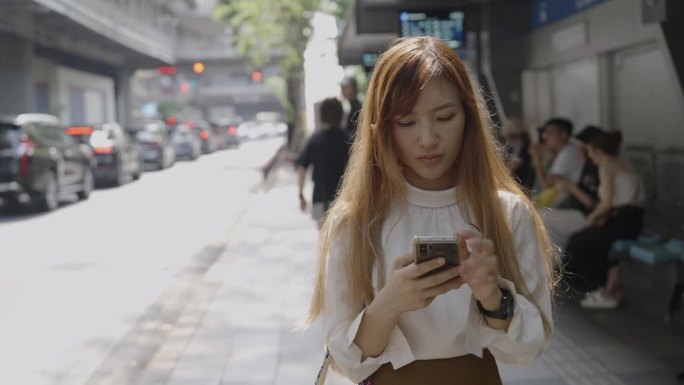 一位亚洲女士站在路边，一边等车来接她，一边刷着手机查看需要多长时间。行人和坐在公共汽车站上的人看起来