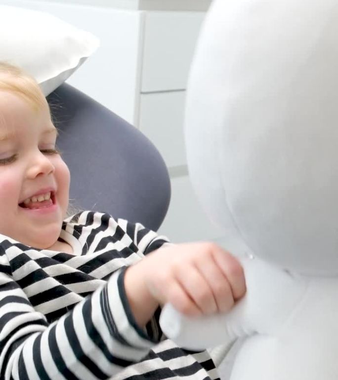一个4.5岁的女孩在牙科椅上玩一个毛绒玩具，一个疯子天线宝宝或一个宇航员，她挥手，它看起来微笑放松等
