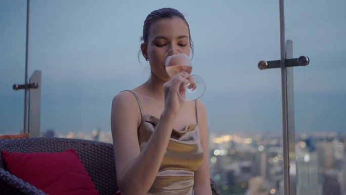 身穿金色礼服的优雅女子在豪华的屋顶酒吧喝着粉红色的葡萄酒。