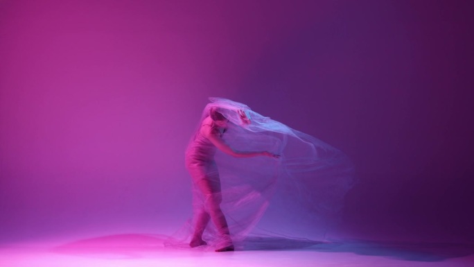 光明和自由。年轻温柔的女孩，芭蕾舞者在霓虹灯的渐变粉色紫色工作室背景下，用透明的面纱跳舞