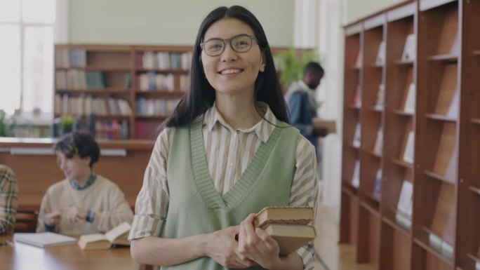 一个无忧无虑的亚洲女孩，戴着眼镜，穿着时髦的衣服，站在大学图书馆里拿着书