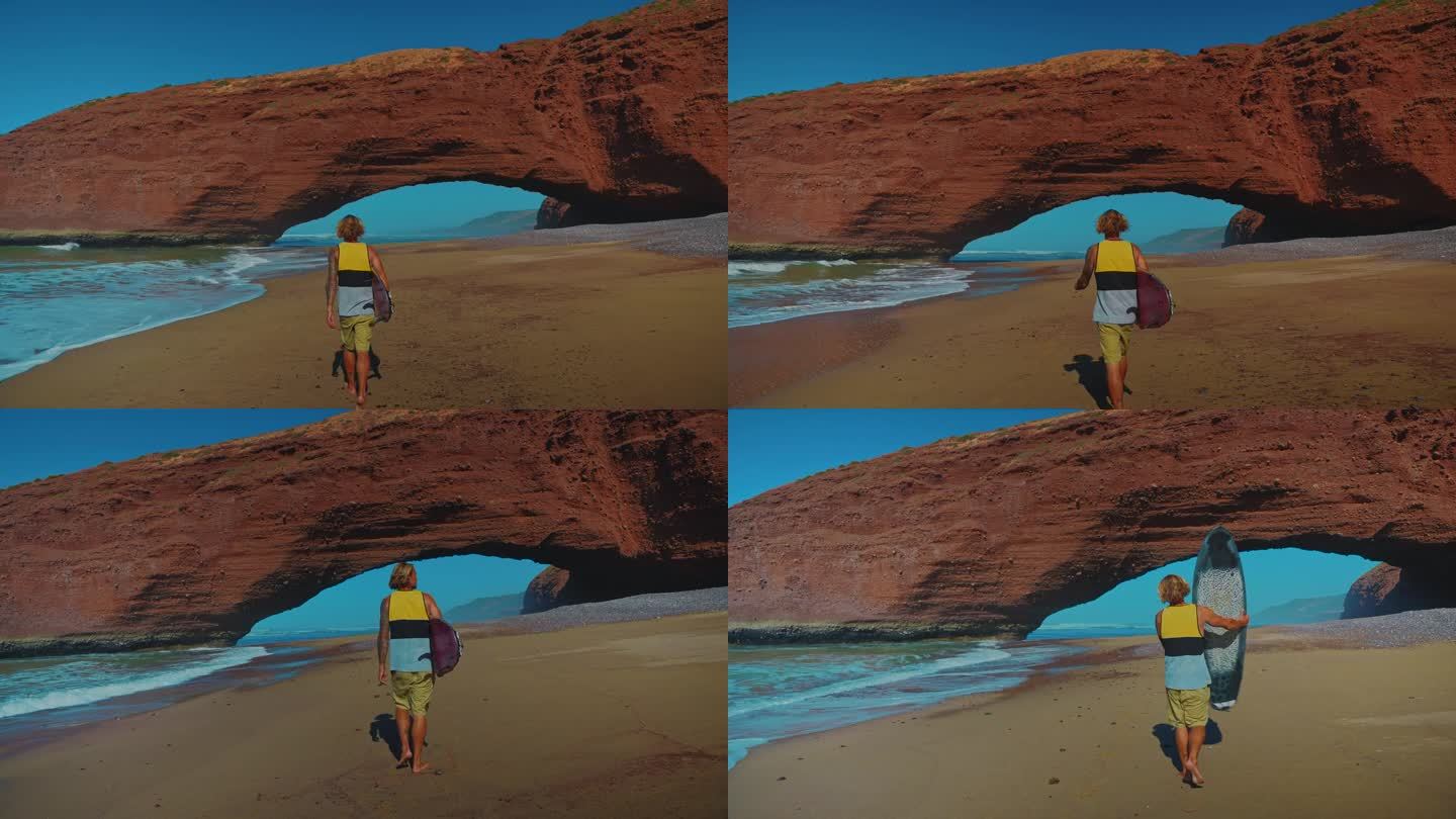 一名男子在沙滩上散步，腋下夹着冲浪板，背景是令人惊叹的岩层