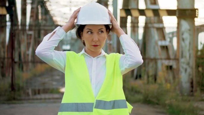 自信的女工程师，穿着工作服，戴着安全帽，站在金属桥建筑附近，在艰苦的工作前戴上头盔