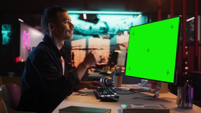 年轻人的肖像坐在他的办公桌上使用台式电脑与模拟绿屏。男性高加索专家工作与彩色键显示在创意机构的电脑