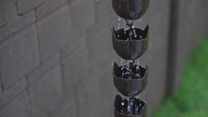 SLO MO:倾斜向下拍摄的装饰钟形金属雨链连接到屋檐上的排水沟，雨水从上到下流下来。