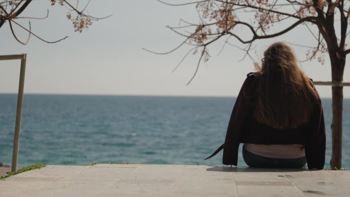 一位年轻女子坐在海边的混凝土上，望着远方。风吹乱了她的长发。