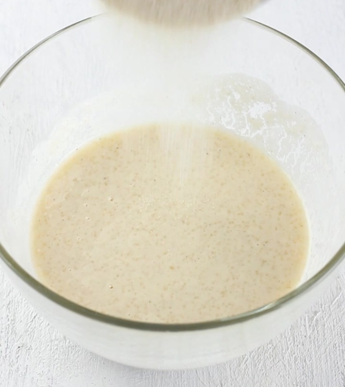 准备面团，面粉蛋糕或馅饼，在浅灰色背景下，将面粉倒入玻璃碗中的蛋奶油混合物中。烹饪阶段，家庭烘焙