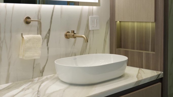 白色水槽的侧视图与金色水龙头在设计师和光线充足的浴室。大理石和木墙与现代家具相辅相成，创造出昂贵的外