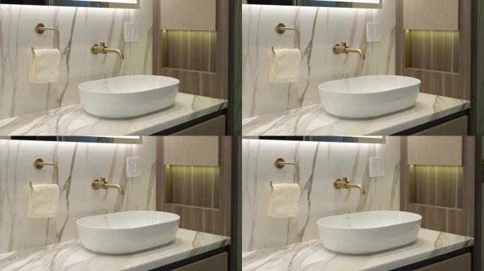 白色水槽的侧视图与金色水龙头在设计师和光线充足的浴室。大理石和木墙与现代家具相辅相成，创造出昂贵的外