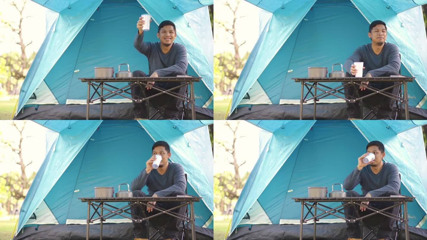 一名亚洲男子坐在露营帐篷前，一边喝着热咖啡，一边在森林露营地休息和放松。
