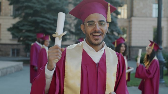 一个快乐的中东人站在大学附近笑着挥舞着文凭指着卷轴的慢动作肖像