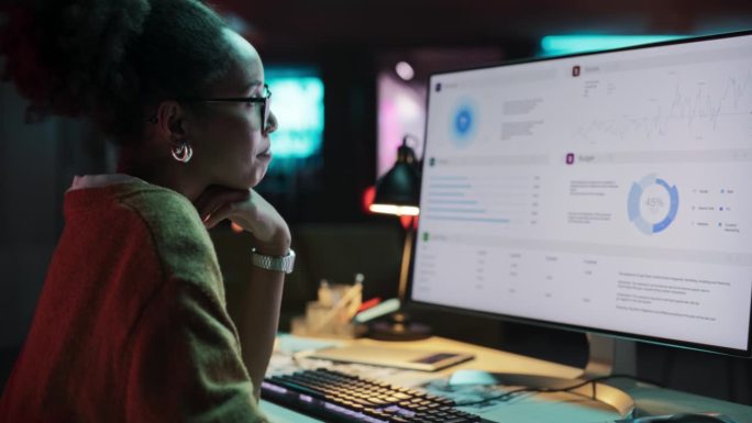 一个快乐成功的黑人女商人的肖像在创意机构使用台式电脑在晚上。非裔美国女性在检查数据和统计图表时微笑