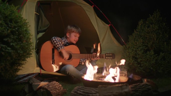 男孩坐在营火旁的帐篷里弹吉他。一个孩子晚上坐在火边唱歌。
