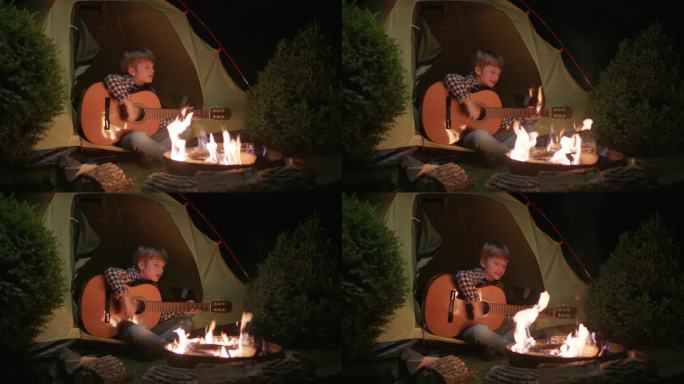 男孩坐在营火旁的帐篷里弹吉他。一个孩子晚上坐在火边唱歌。