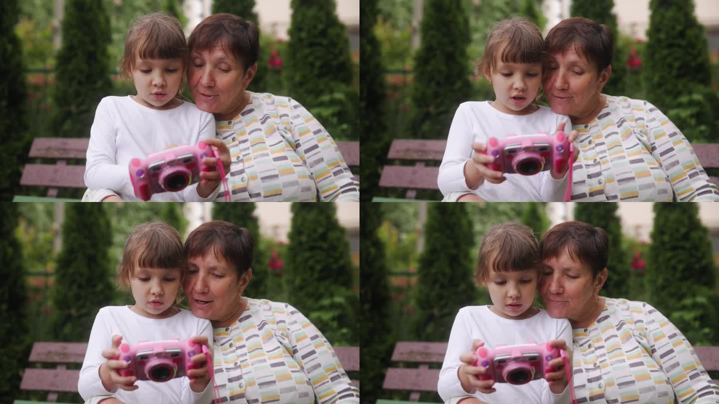 一个甜美美丽的小女孩用她的玩具相机给奶奶看照片。一家人坐在长凳上，老妇人和孩子一起欢天喜地地庆祝。不