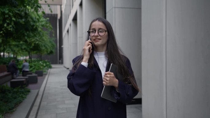 一位留着长发的年轻女子，穿着经典的休闲装，戴着眼镜，走在市中心的办公楼附近，拿着平板电脑，微笑着讲着