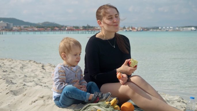 微笑的女人和她年幼的儿子在沙滩上野餐，享受着咸咸的海风和甜甜的苹果。