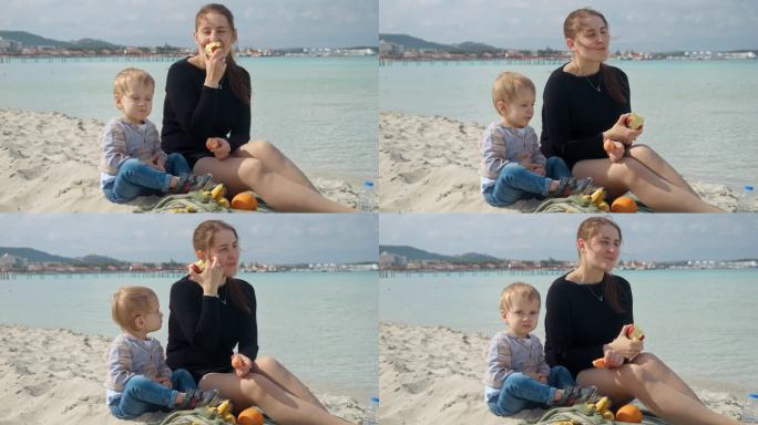 微笑的女人和她年幼的儿子在沙滩上野餐，享受着咸咸的海风和甜甜的苹果。