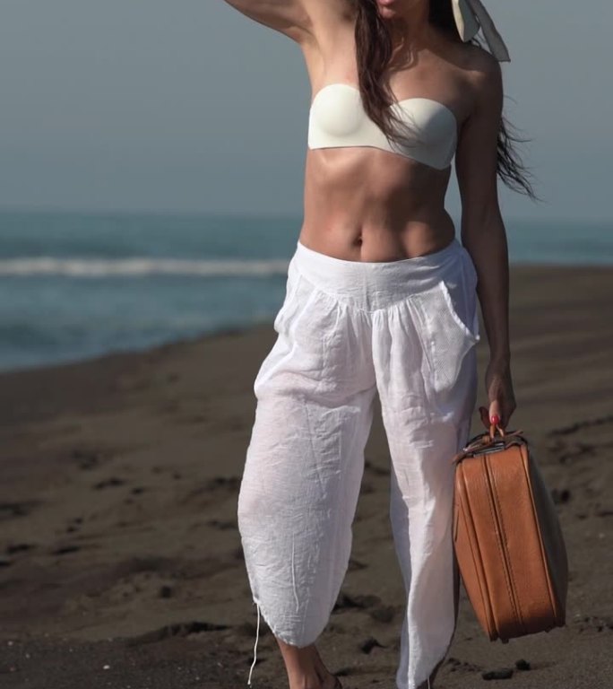 赤脚的女性提着棕色的行李箱站在沙滩上，在夏季的海滩度假时，她望向别处