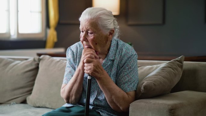 心烦意乱的80年代退休老太太，手里拿着拐杖，一个人沮丧地坐在家里的沙发上
