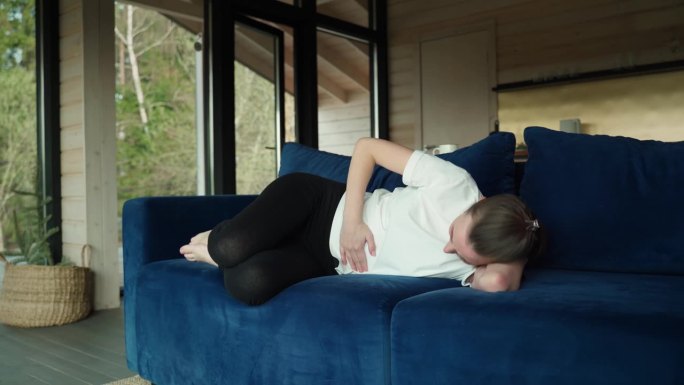 悲伤的年轻女子穿着便服，痛经，胃恶心，抱着肚子，在月经期间腹部痉挛，躺在家里的蓝色沙发上