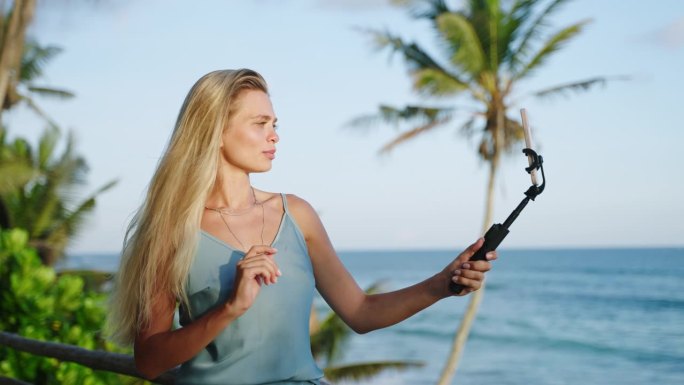 金发美女旅游博主在热带岛屿上用自拍相机拍摄自己的侧视图，可以看到海洋。斯里兰卡一名女视频博主手持手机