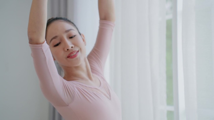 一名身着粉色芭蕾舞裙的优雅女子在舞蹈工作室里举手舞姿，年轻的亚洲女芭蕾舞演员在表演艺术活动中练习芭蕾
