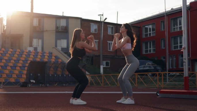 两个女孩，身材苗条的朋友，来到体育场慢跑或健身，做肌肉热身，互相扶持，做双蹲，在体育场进行训练。