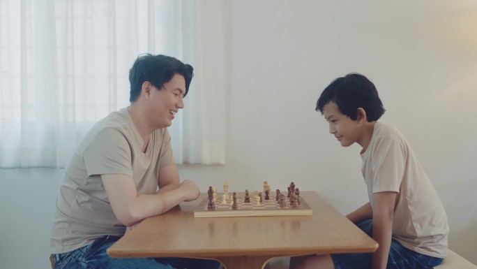 周到英俊的亚洲父亲和可爱的儿子在下棋，而周末家人在家里的房间里休闲和亲密。