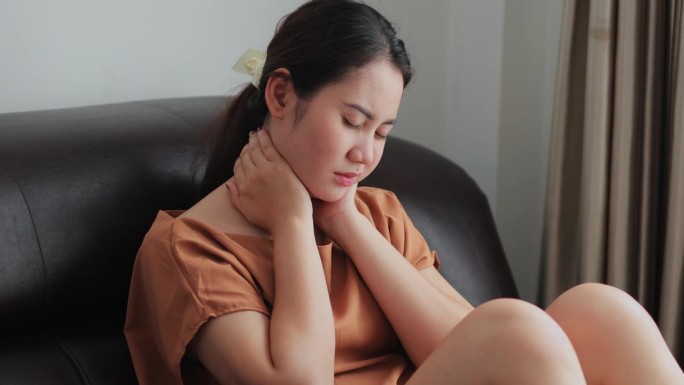 疲惫的亚洲年轻女子因剧烈疼痛而用手摸自己的脖子。在家的沙发上，肌肉遭受纤维肌痛的折磨。