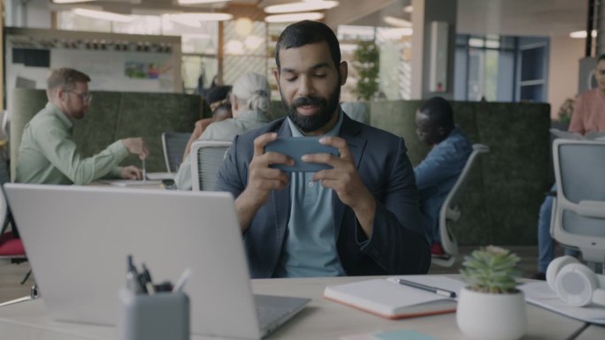 快乐的阿拉伯商人在办公室的办公桌上用智能手机玩在线视频游戏的画像