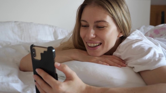 迷人的金发女郎在家里的床上一边讲手机一边开心地笑着。异地恋