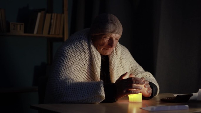 冻僵的老人穿着暖和的夹克，坐在没有中央暖气的寒冷房间里，试图用燃烧的蜡烛加热手掌。没有暖气