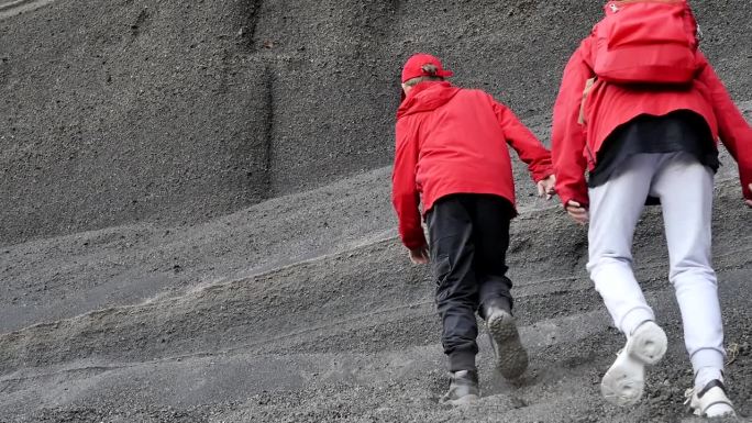 一个小男孩和一个小女孩穿着红色夹克，背着背包，爬上一块由火山灰和古老的死火山渣组成的灰色松散岩石。青