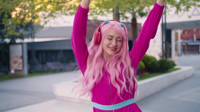 一个穿着粉红色衣服，戴着红色无线耳机的快乐开朗的女孩在街上跳来跳去听音乐。跟着音乐走