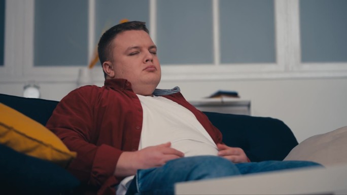 抑郁的肥胖男人坐在沙发上，抚摸着肥胖的肚子，为超重而烦恼