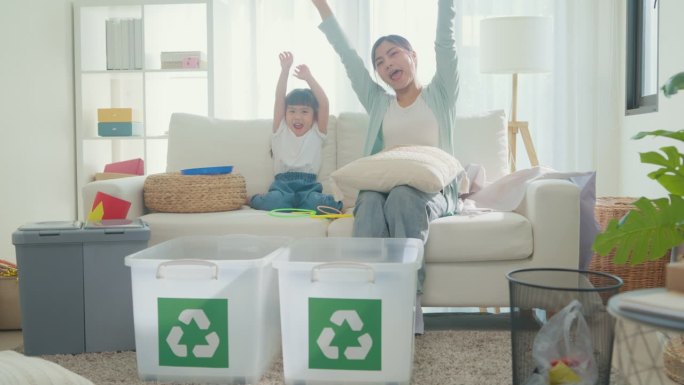 年轻的亚洲家庭妈妈教孩子如何回收，帮助女孩意识到环境的重要性，微笑着看着家里客厅的相机。家庭幸福时刻