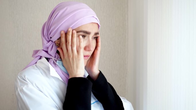 身穿粉色头巾和工作服的穆斯林医生抱着头望着窗外。